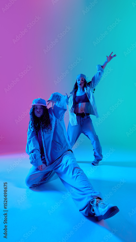Fototapeta premium Artistic teenage girls in denim squatting and dancing hip hop under teal and pink neon lights against gradient studio background. Concept of dance, music, expression, and movement.