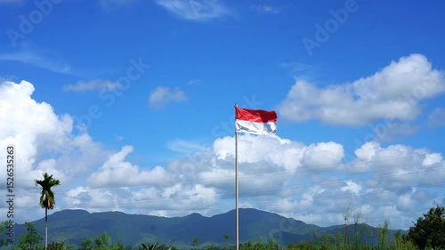 The red and white flag fluttering in the wind on a sunny day. Indonesian flag with blue sky, white clouds and mountains in the background.