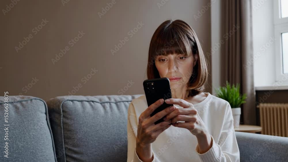 A woman sits on a couch holding a Phone, displaying the screen in the center of the frame, raised to eye level with light brown walls and beige carpet behind her A woman sits on a couch holding a Phone, displaying the screen in the center of the frame, raised to eye level with light brown walls and beige carpet behind her