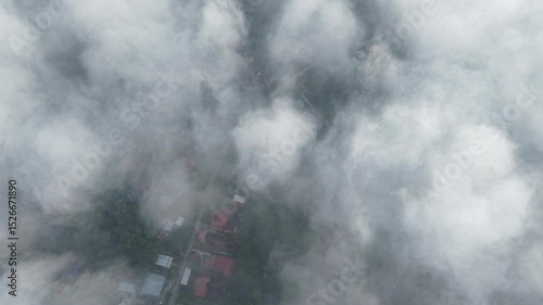Aerial drone footage showcasing a traditional Malaysian village nestled beneath low-hanging clouds. Wooden houses on stilts, winding paths, and lush greenery create a peaceful, nostalgic scene