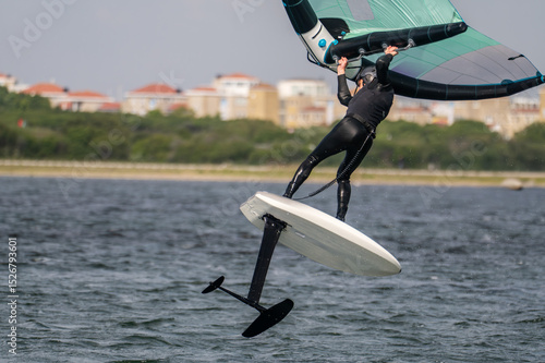 Close-up from a man in a black wet suit jumping with a wing foil at sunny weather in Netherland.