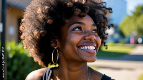 A young woman with curly hair is smiling brightly outdoors on a sunny day.