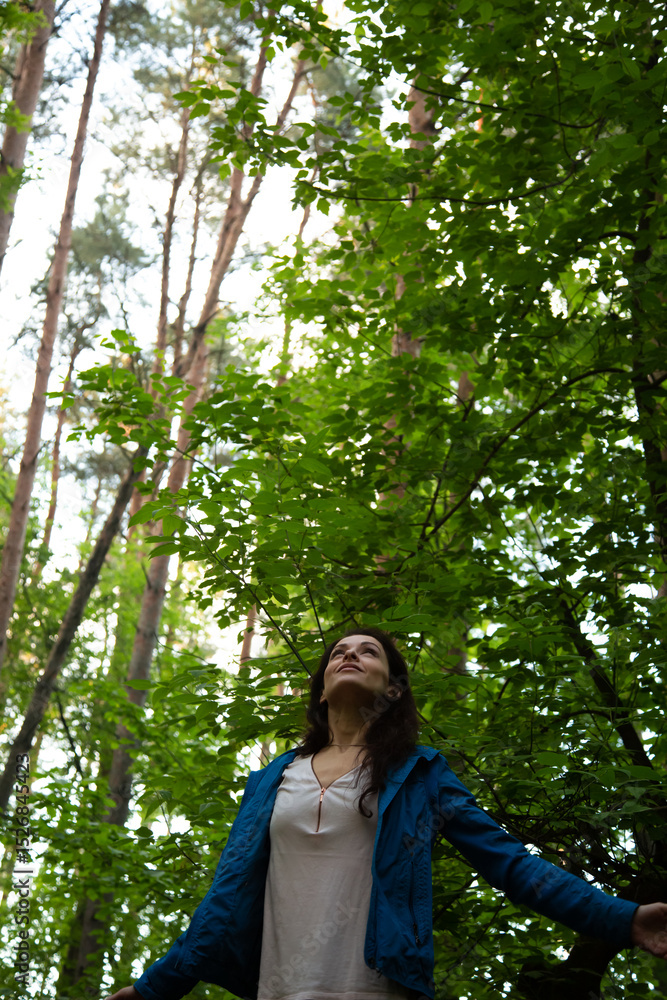 Beauty woman making the arms fly sign, looking up in the pine forest. Female gesture free sign in a park with arms raised back. Girl breath in green wood with trees above