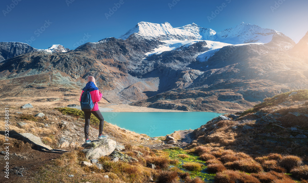 Naklejka premium Young woman hiker with backpack standing on a rock admiring a beautiful turquoise alpine lake with snowy mountain and glacier in the background in fall. Girl on mountain trail in Swiss Alps in autumn
