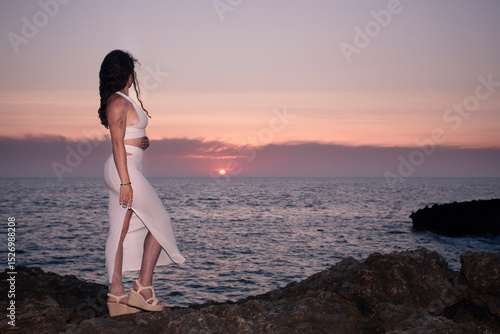 A girl in a white dress watches the sunset from Cap d'Artrutx on the island of Menorca.