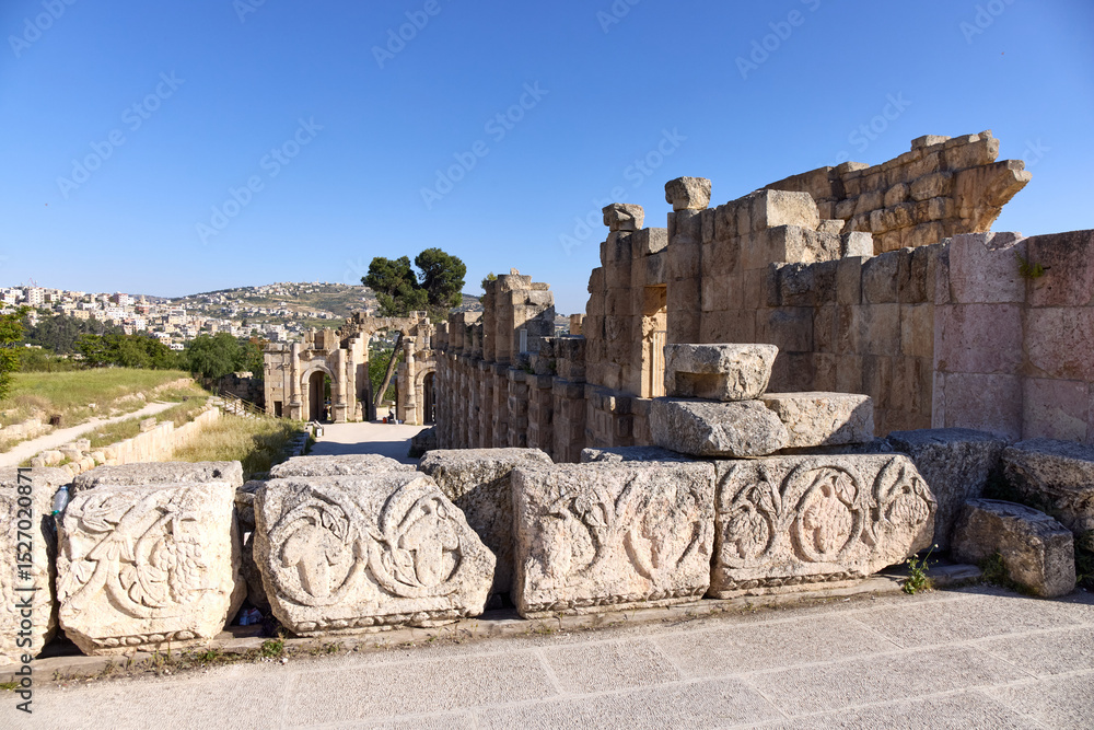 custom made wallpaper toronto digitalVisitors walk among the well-preserved ruins of Jerash, marveling at the intricate carvings and ancient architecture under a bright blue sky, surrounded by lush greenery and distant hills.