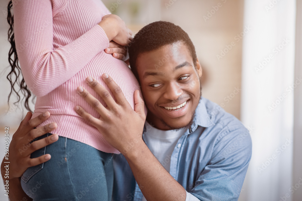 Fototapeta premium Caring black guy putting his ear on girlfriend's pregnant tummy, listening to baby's heartbeat at home. Millennial African American family expecting child, expressing love to each other