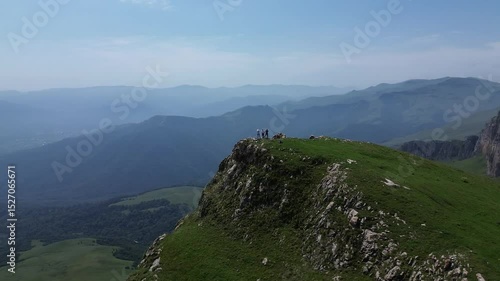Circular drone flight around a rocky hill in green mountains with three people standing on top. Beautiful landscape and terrain set against a clear blue sky.
