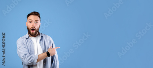 Shocked young guy standing with open mouth, pointing at empty space in disbelief, blue studio background, banner design. Overwhelmed bearded man offering huge sale or promotion