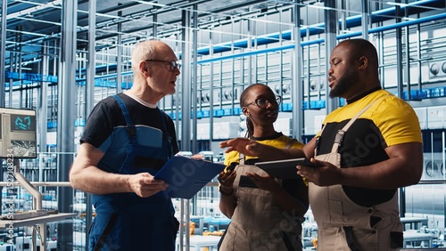 Smart factory engineering team coworkers oversee operation of robotic arms on facility floor. Industry 4.0 plant teamworking colleagues monitor system performance and troubleshoot issues, camera A