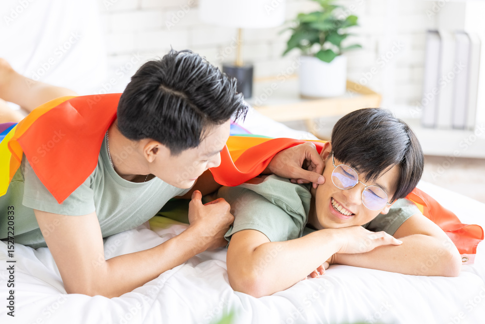 Obraz premium Portrait of carefree gay couple with lgbt flag on the bed, Gay couple love, happy together concept. LGBTQI lifestyles at home.