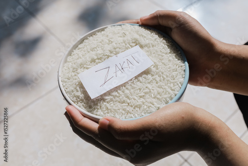 Zakat Rice or zakat fitrah. Rice grains in a glass bowl with zakat written on paper. Islamic concept of zakat to help the poor in need. held by a woman's hand