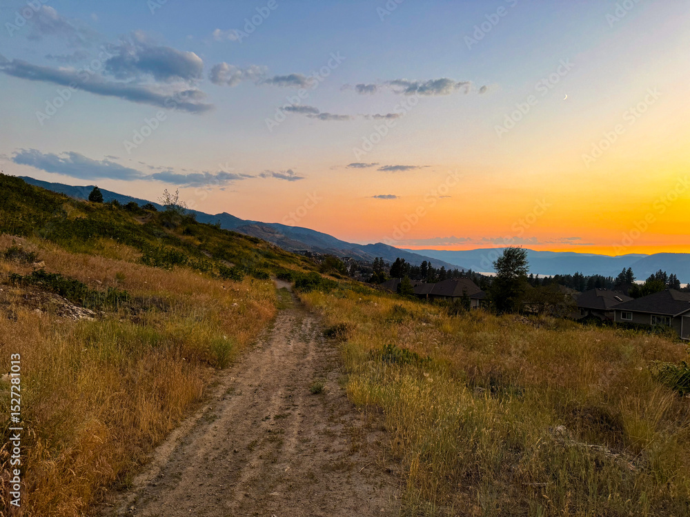 A dirt path winds through sun-kissed fields and hills, leading towards a vibrant sunset painting the sky with warm hues over a distant lake and town. A dirt path winds through sun-kissed fields and hills, leading towards a vibrant sunset painting the sky with warm hues over a distant lake and town.