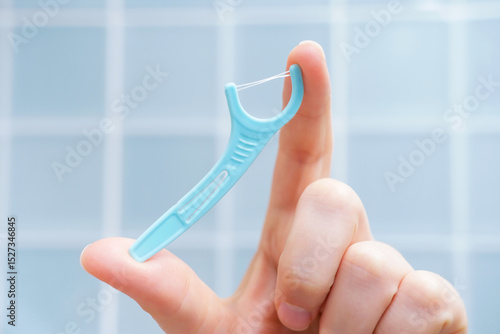Female hand holding disposable dental floss close-up against the background of blue tiles in the bathroom.