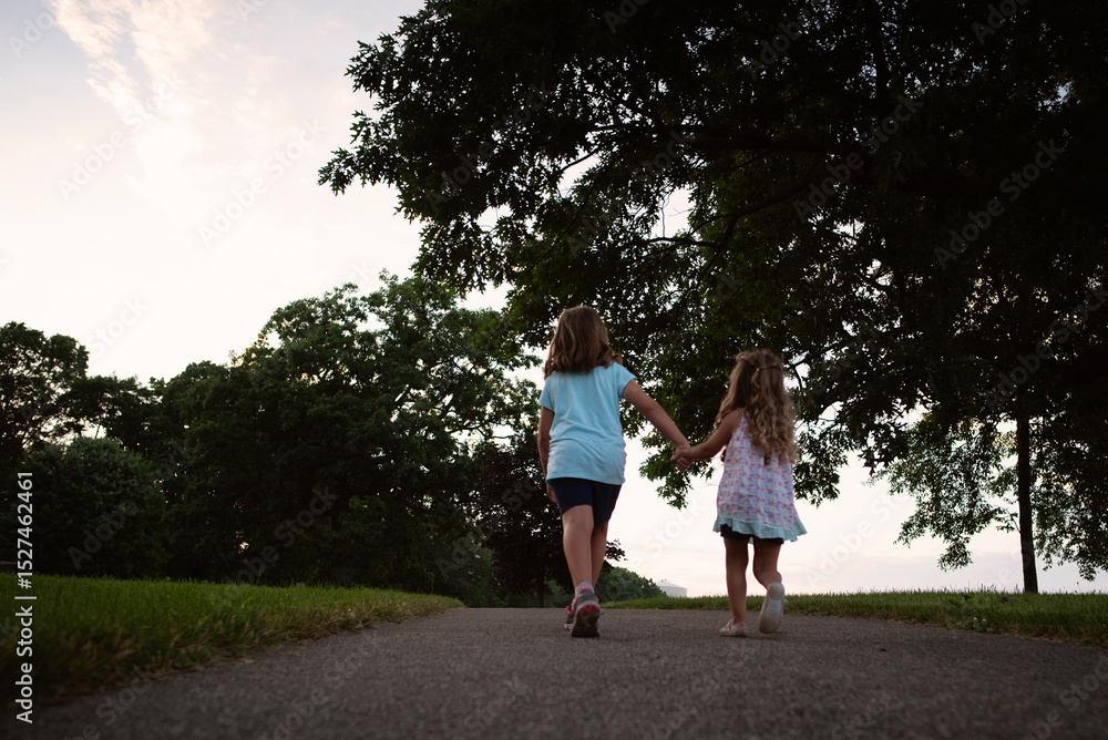 Behind young sisters holding hands walking on path near grassy f Behind young sisters holding hands walking on path near grassy f