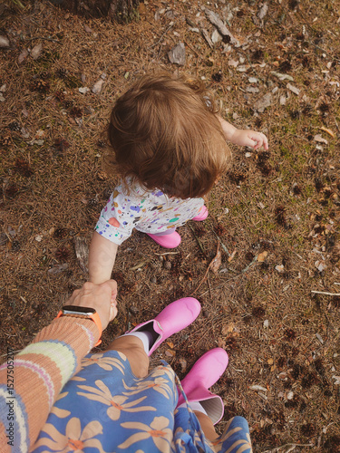 A first-person perspective shot of a walking mother and baby. The toddler is wearing pink rubber boots. The ground is covered in brown pine needles and fallen leaves. Mama is holding daughter's hand. 