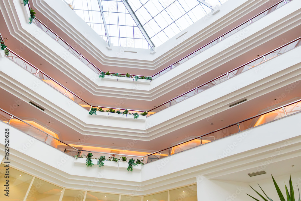 Obraz premium Contemporary hotel atrium with several floors, indoor balconies decorated with plants, and warm ambient lighting. A glass roof allows natural light to enhance the elegant architectural lines.