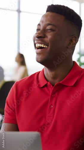 A young black man, late 20s, wearing a vibrant red collared shirt, laughing joyfully with crinkling eyes in a modern, open-plan office bathed in soft natural daylight, with blurred colleagues and
