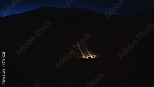 A car travels down a winding mountain road at night, its headlights illuminating the dark path ahead. The scene evokes a sense of adventure and the thrill of alpine driving. Aerial view.