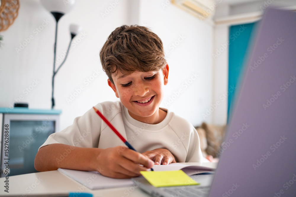 Young boy happily doing homework and writing notes at home while studying with the laptop. He's on a bright living room during the afternoon. Young boy happily doing homework and writing notes at home while studying with the laptop. He's on a bright living room during the afternoon.