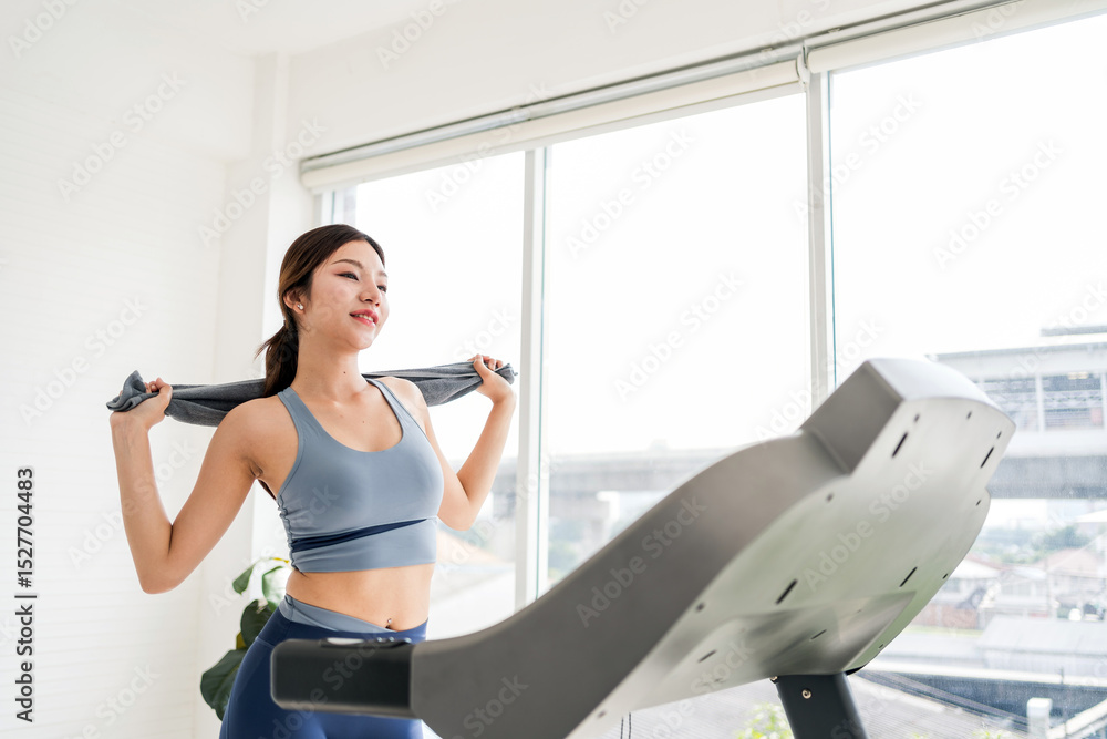 Fototapeta premium Confident young woman exercising indoors on a treadmill, smiling post-workout while holding a towel. Captures healthy lifestyle, fitness motivation, and wellness in a bright home gym setting.