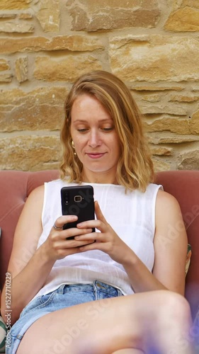 
Woman sitting on pink couch looking at phone with slight smile, static camera with warm natural lighting, empty space in upper left on stone wall background
