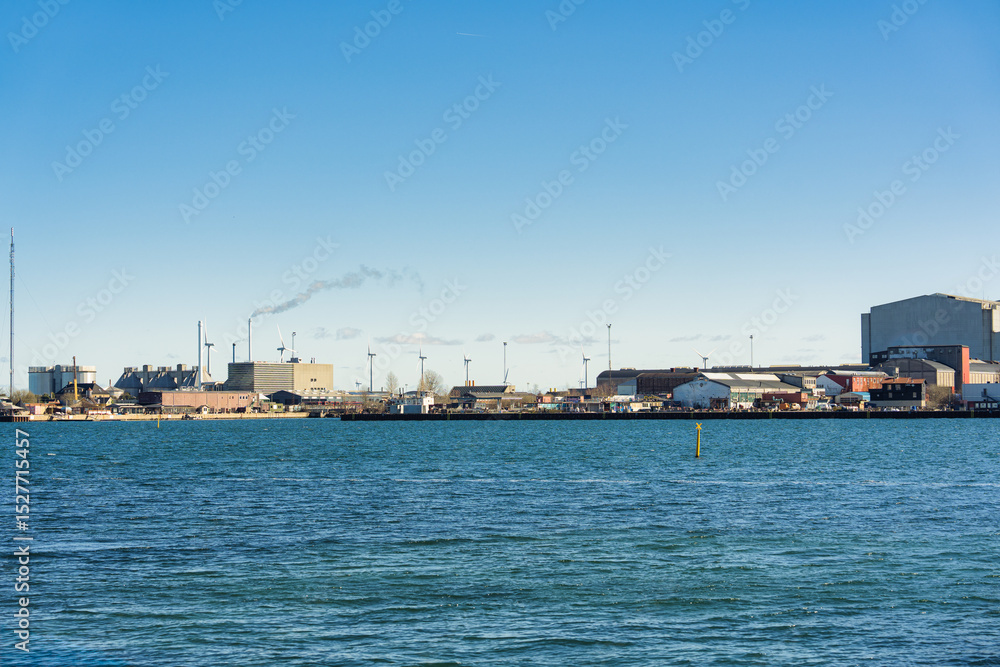 Industrail waterfront with factory building, wind turbine and smoke emitted from chimney on sunny day Industrail waterfront with factory building, wind turbine and smoke emitted from chimney on sunny day