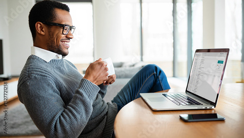 Smiling man relaxing at home, drinking coffee and checking emails on his laptop