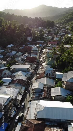 Sweeping aerial video panning a bustling tropical coastal town: dense buildings, winding roads with local people & motorbikes, lush palm trees, and jungle-covered hills under golden hour light,