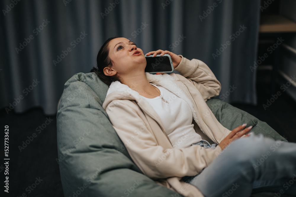 Fototapeta premium Casual scene of a young woman reclining comfortably on a bean bag while talking on her phone, exuding relaxation and contentment in a cozy indoor setting.