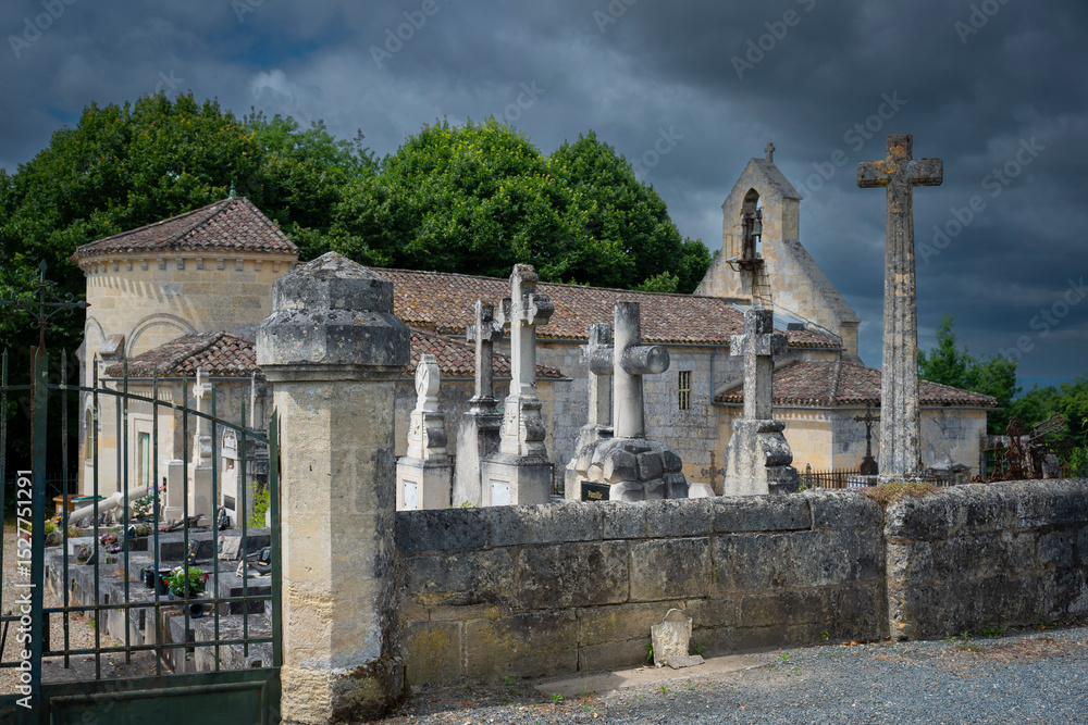 Fototapeta premium A cemetery filled with ornate stone crosses stands adjacent to a historic church in Saint-Laurent-des-Combes, surrounded by lush greenery under a dramatic sky.