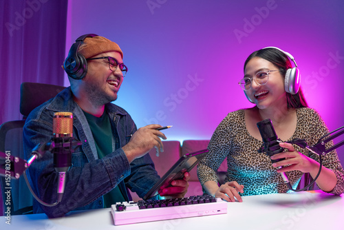 Two young asian streamer host making live podcast with condenser microphone and tablet at broadcast home studio. Content creator and social media influencer creating digital content.