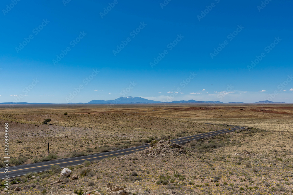 A winding desert road cuts through the vast, barren landscape near Meteor Crater in Winslow, Arizona, with distant mountains under a clear blue sky. A winding desert road cuts through the vast, barren landscape near Meteor Crater in Winslow, Arizona, with distant mountains under a clear blue sky.