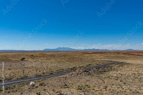 Konstfotografi A winding desert road cuts through the vast, barren landscape near Meteor Crater in Winslow, Arizona, with distant mountains under a clear blue sky