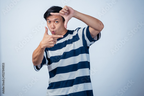 Asian man in striped shirt playfully framing his eye with fingers, pretending to take a photo, smiling against a plain background