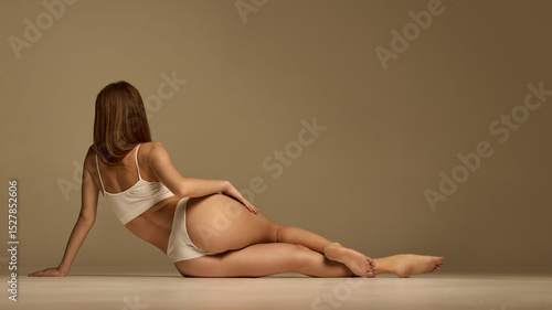 Photography Back view of woman reclining on arm with hand on hip, smooth curves and toned legs emphasized by relaxed pose against beige background