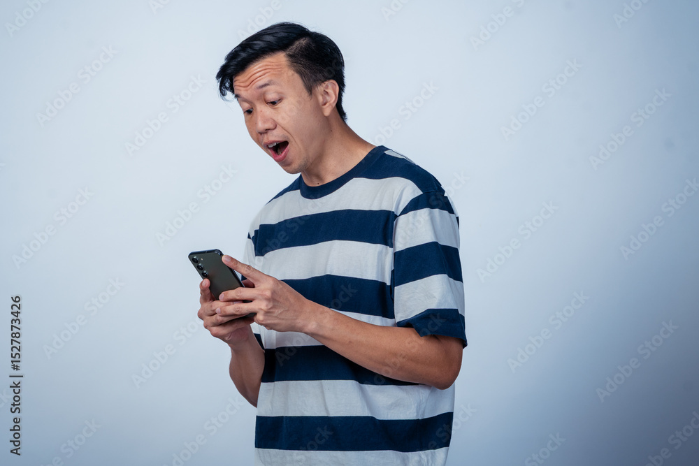Fototapeta premium Asian man in a striped shirt holding a smartphone with a surprised and playful expression, looking directly at the camera against a plain background