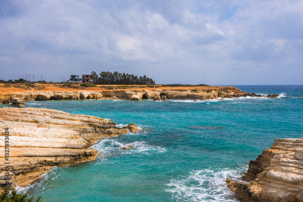 Obraz premium Rugged limestone cliffs meet turquoise waters in Cyprus, with unique rock formations, small sea caves, scattered buildings, and a partly cloudy sky.