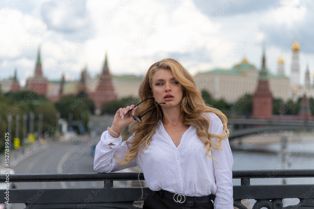 Fototapeta premium Portrait of beautiful blond long haired business woman in eyeglasses, white shirt and black skirt standing on bridge in downtown city district. Moscow Kremlin in the background. Business travel theme.