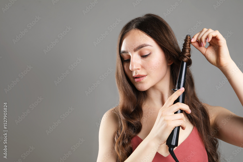 Fototapeta premium Beautiful woman curling her hair on grey background. Space for text