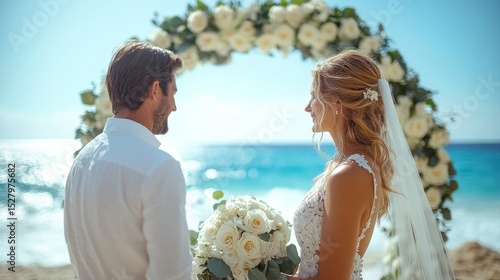 Couple stands facing each other by the shoreline, surrounded by a floral arch while exchanging vows. The ocean waves sparkle under a clear blue sky as they embrace their special moment.