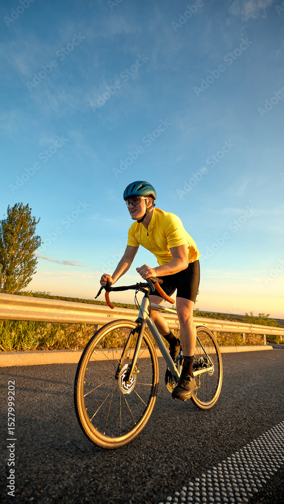Fototapeta premium Focused cyclist sprints through final stretch of training route at golden hour with low sunlight reflecting off asphalt and sky turning orange. Concept of sport, cycling, racing, training, endurance