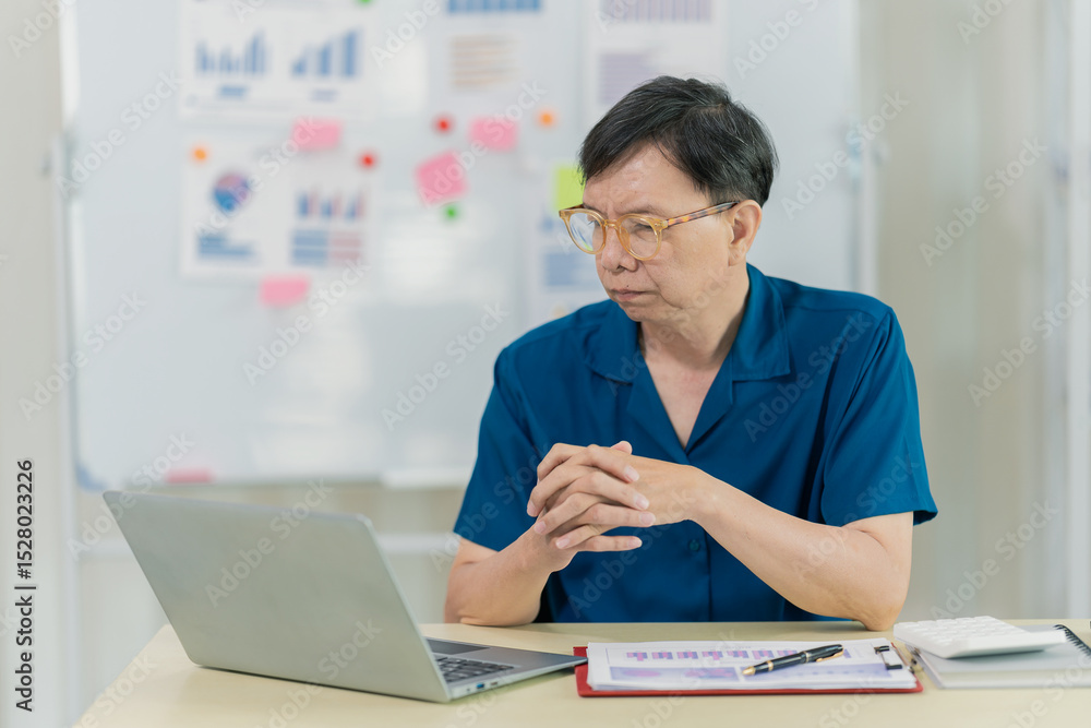 Fototapeta premium Asian man sitting at desk in front of laptop at home preparing budget and financial documents related to household expenses.