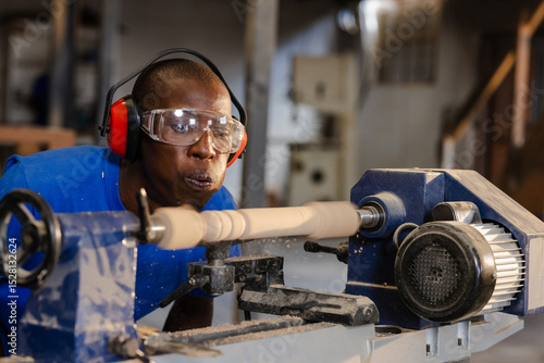 African American man in coveralls turning lathe spindle in workshop blowing shavings, copy space