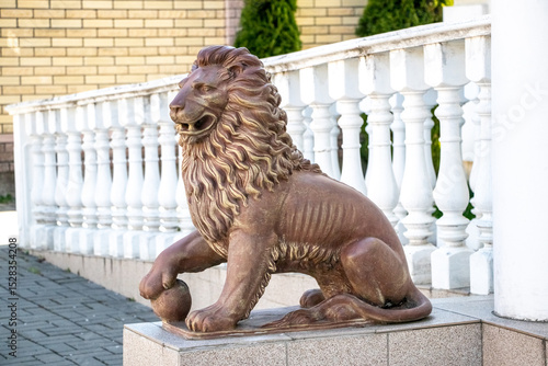 Fototapeta A stone lion statue with a mane, seated on a pedestal near white balusters and a