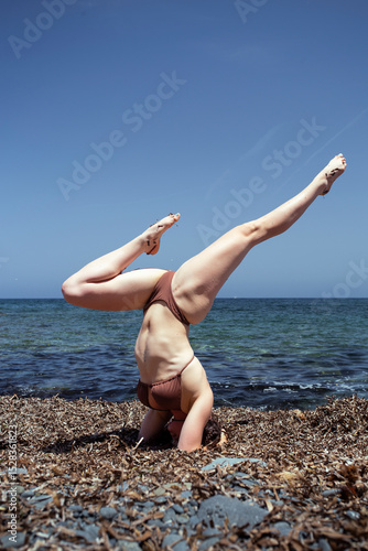 Young girl in bikini performs an inverted yoga pose on a blanket of Posidonia oceanica on the beach