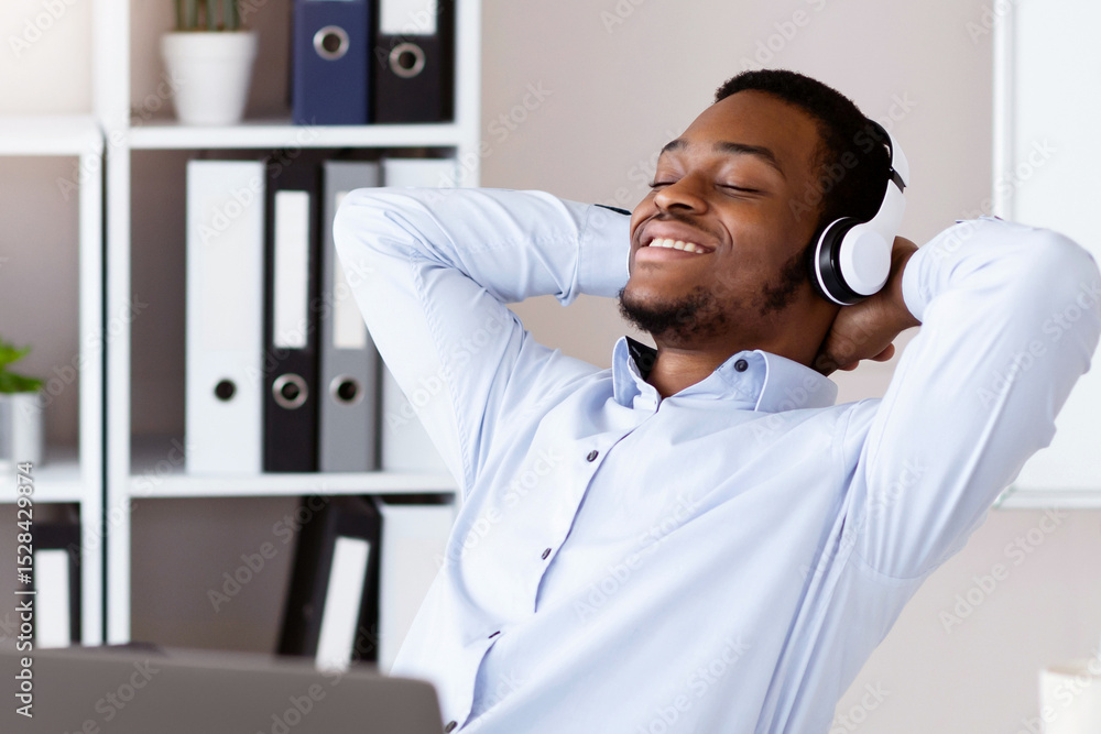 Peaceful black manager leaning back with legs on workdesk and listening to music with closed eyes in his office, using laptop and wireless headset, copy space. Stress relief at job concept Peaceful black manager leaning back with legs on workdesk and listening to music with closed eyes in his office, using laptop and wireless headset, copy space. Stress relief at job concept