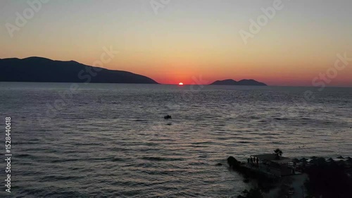 Upward drone footage capturing a tranquil boat gliding over the Ionian Sea during sunset, with the silhouette of Karaburun Peninsula framing the horizon in southern Albania.