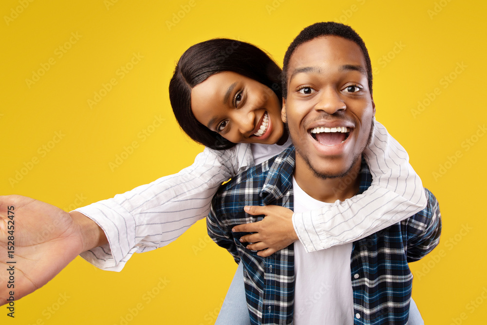 Fototapeta premium Portrait of lovers on date and have fun together. Happy young african american male holds woman on back, laughs with open mouth, lady makes selfie, isolated on yellow background, studio shot