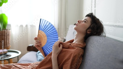 Exhausted woman lying on sofa soaked in sweat using fan to relieve extreme heat. Heatstroke risk, indoor climate discomfort, physical fatigue, urban summer survival, dehydration, no conditioning